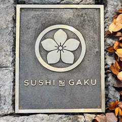 Bronze address sign with 'Sushi Gaku' and a cherry blossom design on a stone wall.