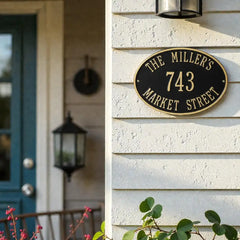 Oval bronze address sign house number sign on a house exterior with a blue door and greenery.