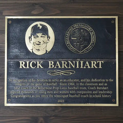 A bronze dedication plaque honoring Rick Barnhiart for his contributions to baseball education, displayed on a wooden surface.