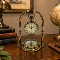 A vintage-style brass mantel clock with a suspended pocket-watch face and a compass base, displayed on a wooden desk with books, a globe, and a lamp in the background.