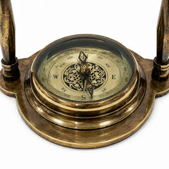 An antique brass compass with an ornate circular dial, glass cover, and two vertical brass legs, shown on a white background.