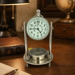 A vintage-style metal desk clock with a round white face, black numerals, and a small compass on the base, displayed on a wooden desk with books and a lamp in the background.