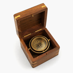 A vintage brass compass displayed inside an open wooden presentation box with brass corner accents, set against a plain light gray background.