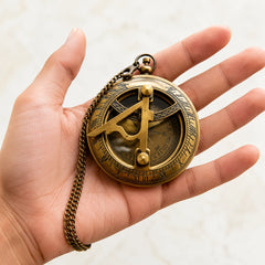 A brass pocket watch designed to look like an antique astrolabe or compass, held in the palm of a hand. The watch features a bronze chain and a detailed brass face with Roman numerals and geometric instruments.
