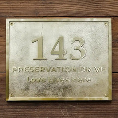 Brass house number plaque engraved | Metallic house number plaque with '143 Preservation Drive Love Lives Here' on a wooden background