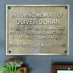 Engraved remembrance plaque in brass | Memorial plaque for Oliver Doran on a gray wall with a plant in the corner.