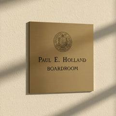 Brass plaque with personalized engraving with 'Paul E. Holland Boardroom' text and university emblem on a beige wall.