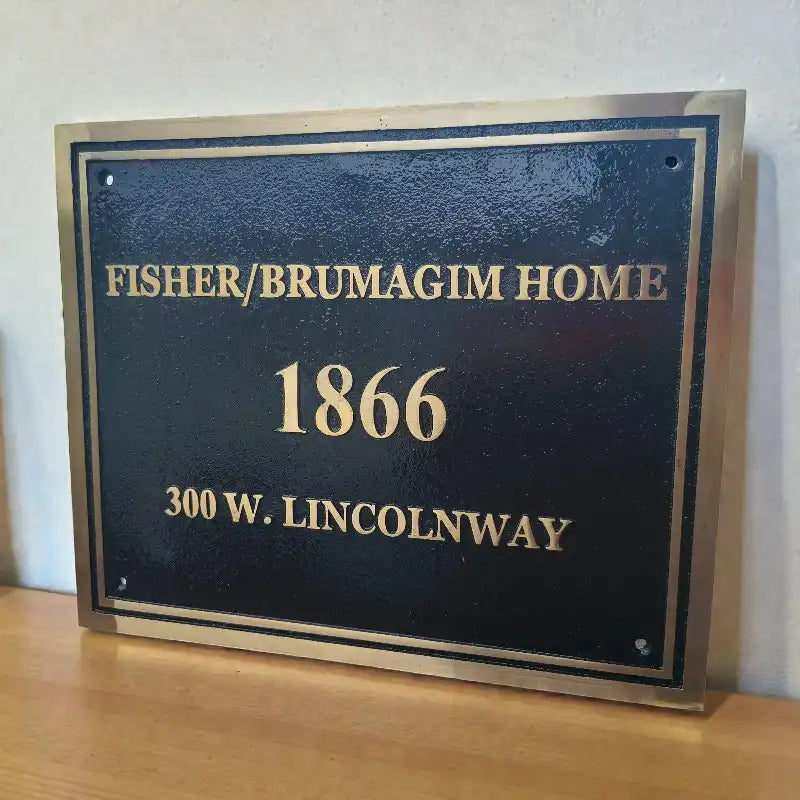 Plaque with text 'Fisher/Brumagim Home 1866 300 W. Lincolnway' on a wooden surface.