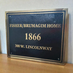 Plaque with text 'Fisher/Brumagim Home 1866 300 W. Lincolnway' on a wooden surface.