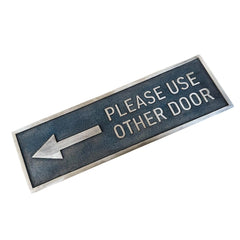 Rectangular bronze door sign with text 'Please use other door' and an arrow on a white background