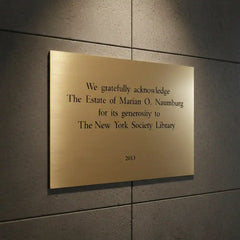Custom Engraved Brass Plaque Front View—A gold plaque on a gray wall acknowledging the Estate of Marian O. Naumburg for its generosity to the New York Society Library.