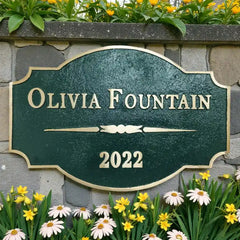 Personalized brass address plaque sign with 'OLIVIA FOUNTAIN 2022' on a stone wall with flowers.