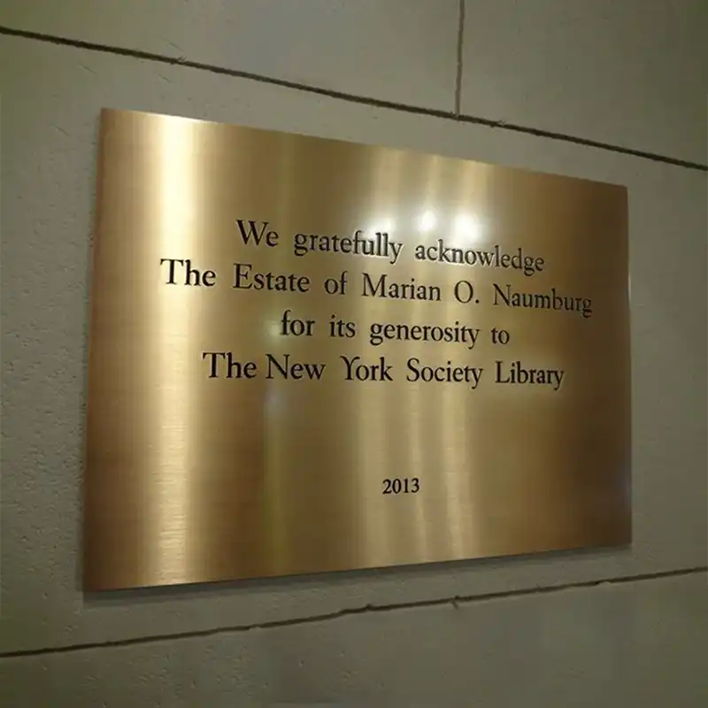 Custom Engraved Brass Plaque Front View—A gold plaque on a gray wall acknowledging the Estate of Marian O. Naumburg for its generosity to the New York Society Library.