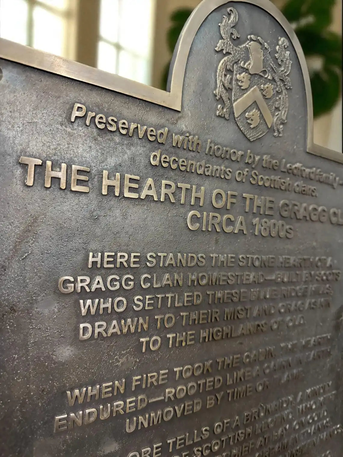 Bronze plaque with engraved text and family crest, set against a blurred indoor background.