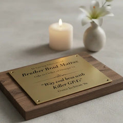 Personalized brass tribute plate on a wooden base with engraved text, next to a lit candle and white flowers.
